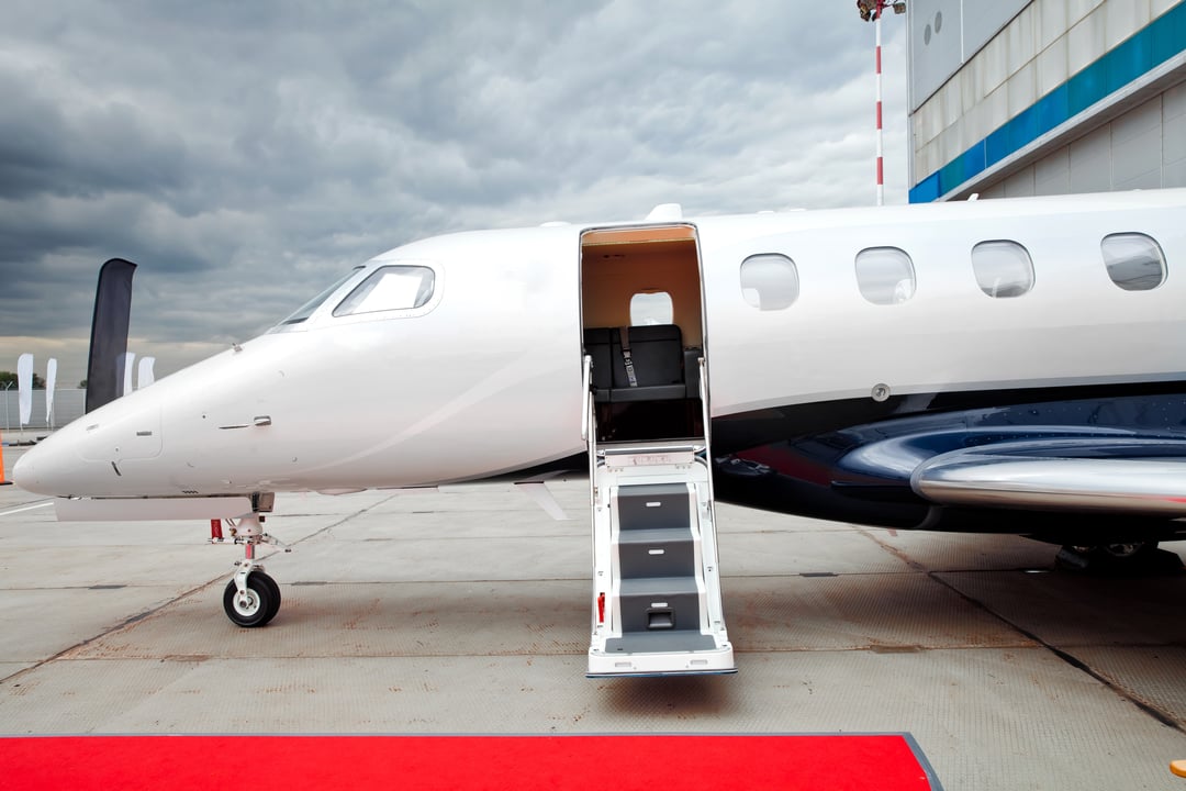Close-up view of a modern private jet with open cabin door and stairs extended onto a red carpet at an airport ramp, under cloudy skies.